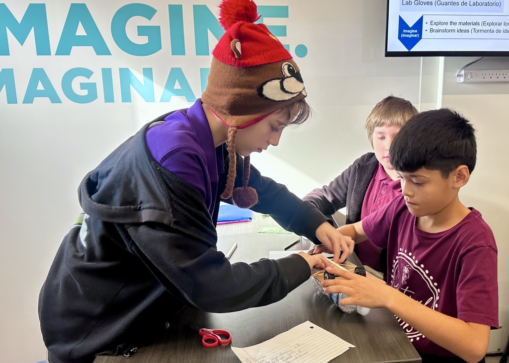 Three elementary boys work together at a table inside a mobile STEM lab, carefully assembling a small bottle rocket truck. One student wearing a knit hat adjusts materials on the bottle while the others watch and assist, with scissors and notes on the table and a STEM design prompt visible in the background.