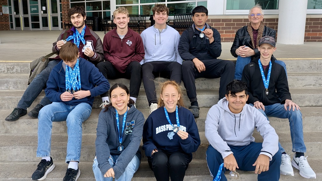 Group of Athens High School students and their coach sitting on outdoor steps, smiling and holding medals from the Mabank Winter Invitational. The students are wearing casual clothes and displaying blue lanyards with multiple medals.