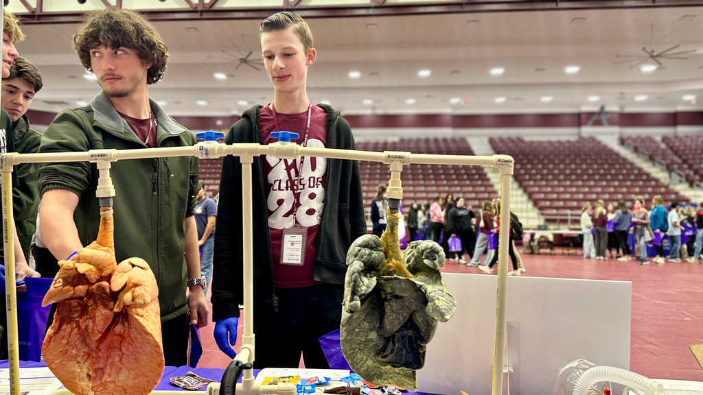 High school students stand at a display table examining two real human lungs hanging side by side, one pink and healthy and the other dark and damaged from smoking, during a health science career fair in a large gym. Students in maroon shirts and gloves look closely at the comparison while other attendees move through the background.
