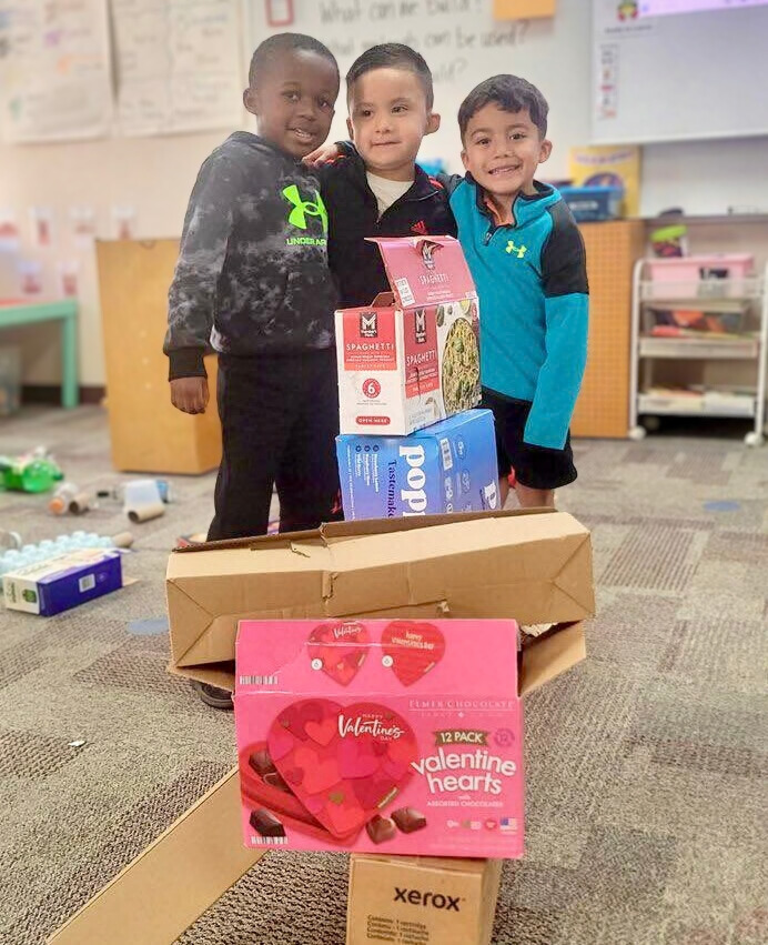 Three young boys stand with arms on each others' shoulders in front of empty cardboard boxes stacked atop one another.