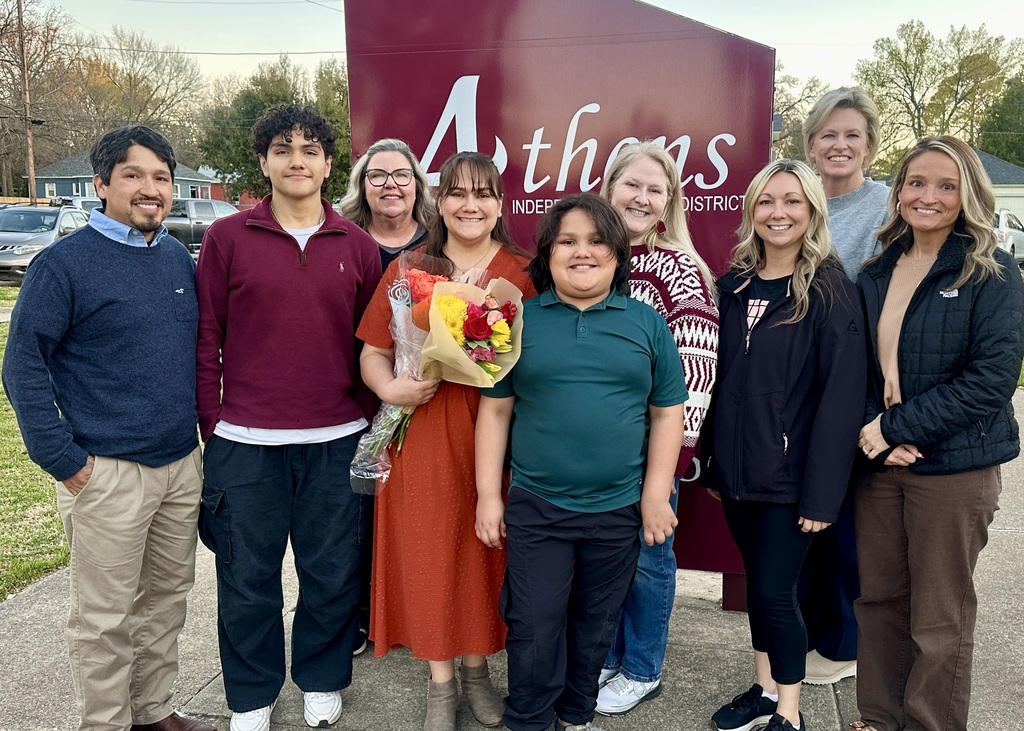 Nine people, including adults and two students, stand together outdoors in front of a maroon Athens Independent School District sign. A woman in the center holds a bouquet of red and yellow flowers, while a student in a green polo shirt stands in front smiling. The group is dressed in casual and business casual attire and appears to be celebrating a recognition or special achievement.