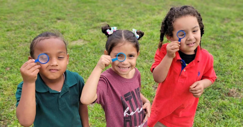 Three young children stand in what appears to be a grassy field or playground peering through plastic magnifying glasses.