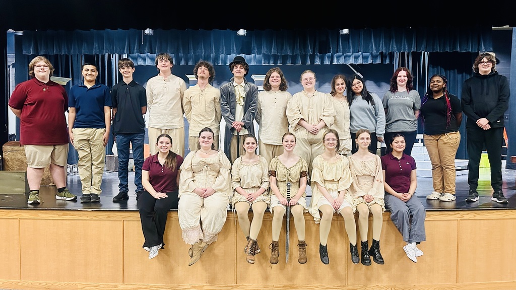A large group of high school students—cast and crew of a one-act play—pose together on a stage. The front row sits along the edge of the stage, mostly wearing coordinated beige, period-style costumes, while the back row stands behind them in a mix of costumes and casual clothing. A student in the center wears a more elaborate outfit with a coat and hat, suggesting a lead role. The stage backdrop features blue curtains and set pieces, indicating a theatrical production setting. The group smiles and faces the camera, suggesting a celebratory cast photo after or before a performance.