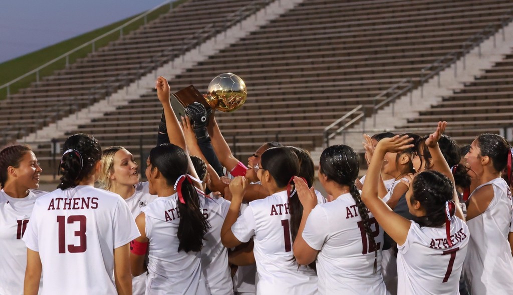 Girls soccer players in white jersies celebrating and holding up a soccer-ball shaped trophy with stadium seats in the background.