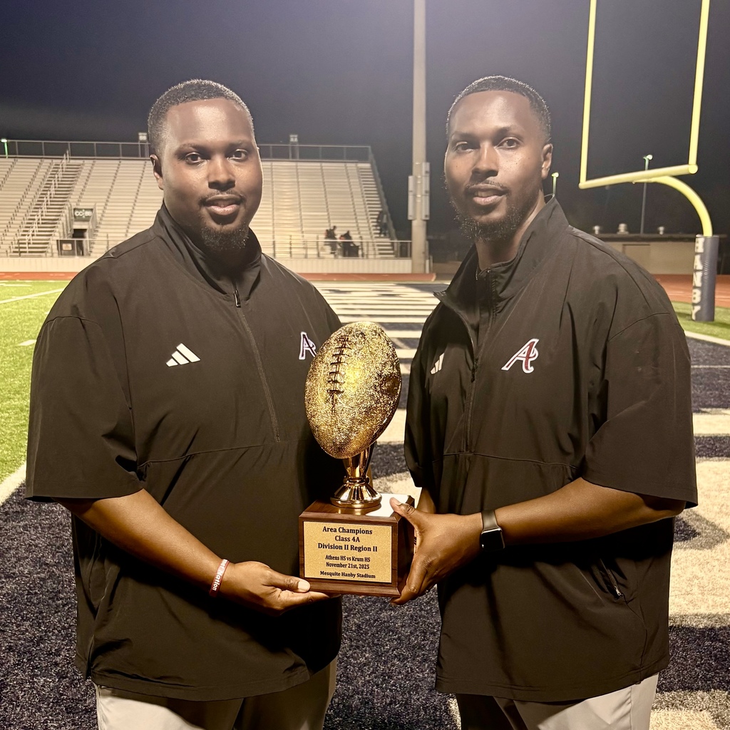 Two adult men stand side by side on a football field at night, holding a gold football-shaped trophy together. They are wearing black athletic jackets with an “A” logo, and stadium bleachers and goalposts are visible in the background.