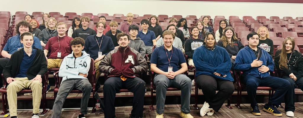 A large group of high school students sit together in rows of maroon auditorium seats, smiling at the camera for a team photo, with several students wearing Athens ISD lanyards and one student in the front row wearing a maroon letterman jacket with an “A.”