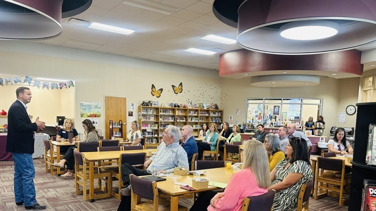 a man in a suit jacket speaks to several professionally dressed people seated around tables inside a library  