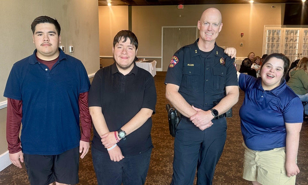 Four people stand together indoors at a community event in a banquet hall. Three young individuals in casual polo shirts and shorts pose beside the Athens Police Chief, who is wearing a dark police uniform and smiling with his hands clasped. One of the youths has an arm around the chief’s shoulder. Tables and other attendees are visible in the softly lit background.
