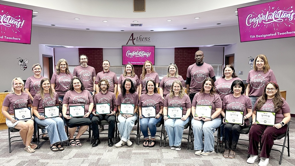 Alt text: Group photo of Athens ISD TIA designated teachers in a district meeting room. Ten educators are seated in the front row holding certificates, with 11 more standing behind them. All are wearing matching mauve Athens ISD T-shirts, and three pink screens in the background display a congratulatory message for TIA designated teachers.