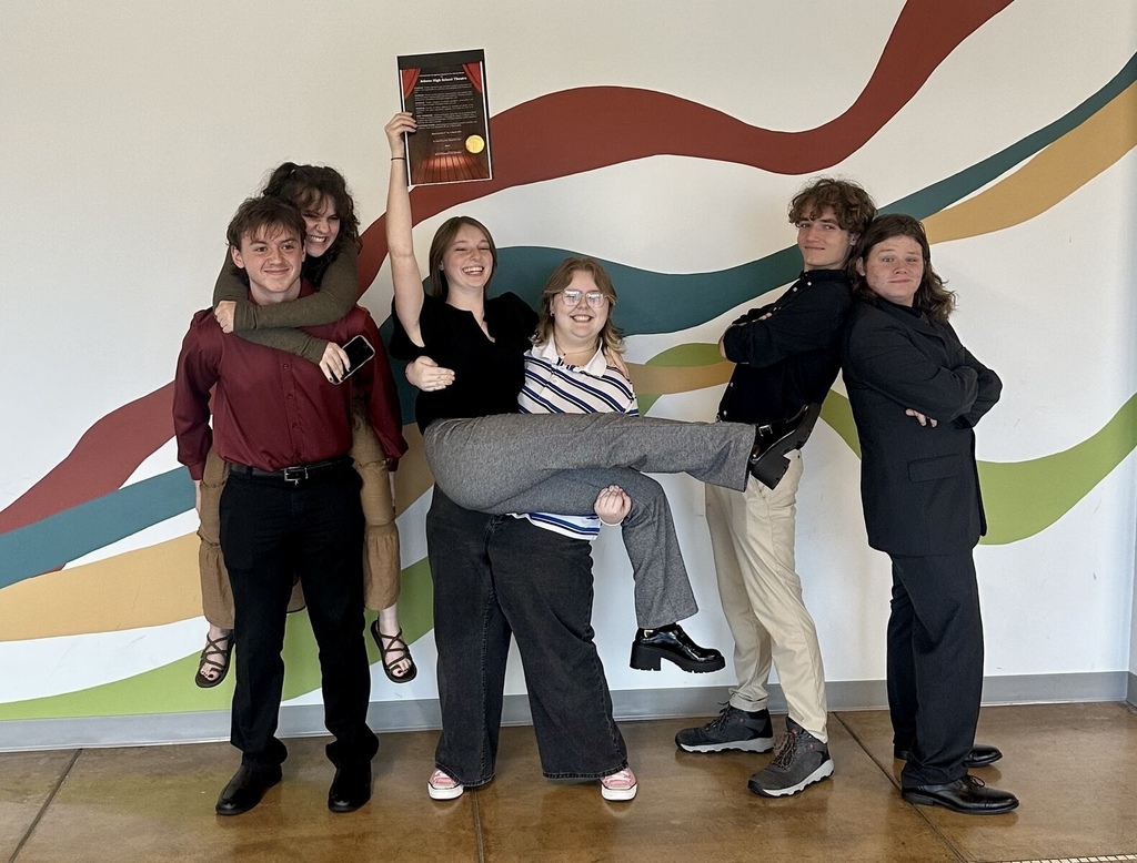 Six students pose playfully in front of a colorful mural, smiling and celebrating as one student holds up a plaque overhead. Two students stand back-to-back with their arms crossed, while others strike fun poses, including one student being held up and another riding piggyback.
