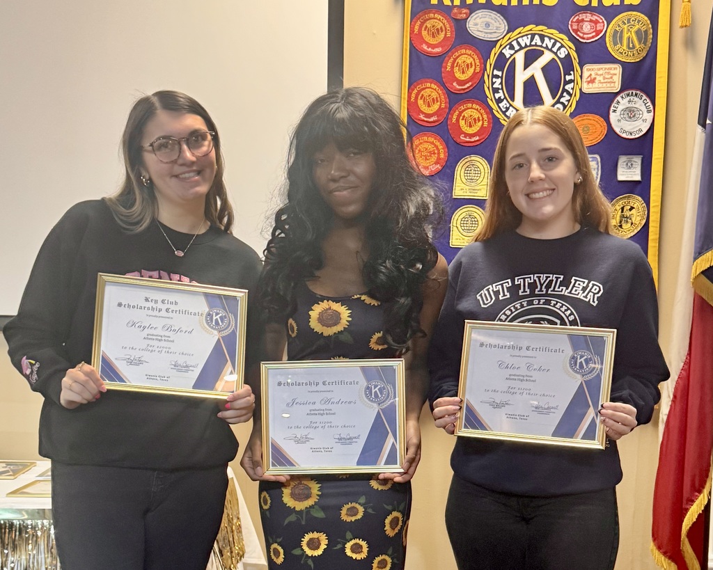 Three young ladies holding framed scholarship certificates standing in front of what appears to be a Kiwanis banner.