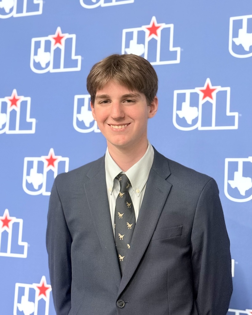 Alt text: A smiling male student wearing a navy suit, light-colored dress shirt, and patterned tie stands in front of a blue UIL backdrop decorated with Texas UIL logos.