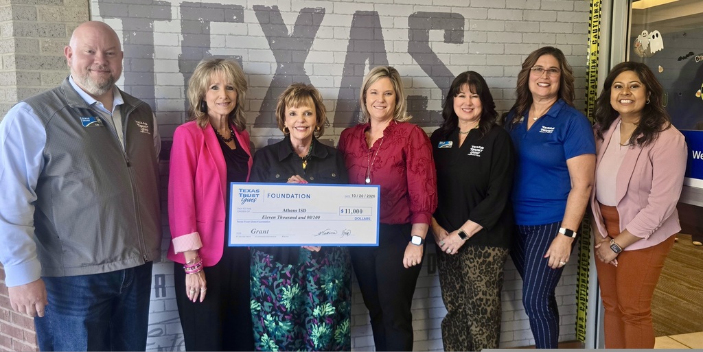 even people stand together for a check presentation from the Texas Trust Gives Foundation to Athens ISD. Three people in the center hold a large ceremonial check for $11,000, dated Oct. 20, 2026. Pictured from left are Cody Smith, Dr. Janie Sims, Ginger Morrison, Brooke Brock, Kristi Hammond, Stacey Barbosa and Washima Huq. The group stands in front of a brick wall with large letters spelling “TEXAS.”