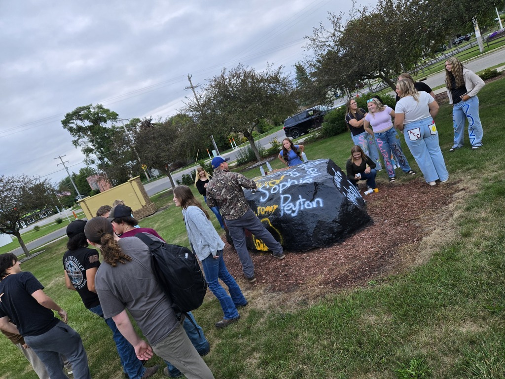 A group of people gather outdoors near a large rock painted with the names “Sophie” and “Peyton” in yellow and blue letters on a grassy area.