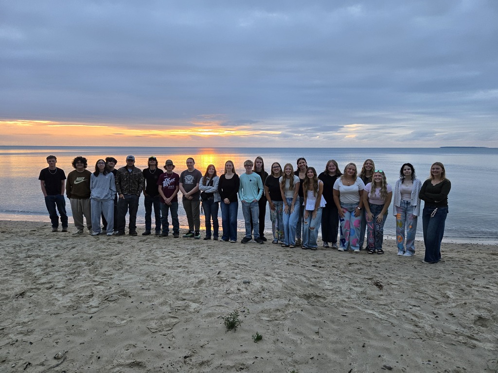 A group of people stand in a line on a sandy beach at sunset, with calm water and a cloudy sky in the background.