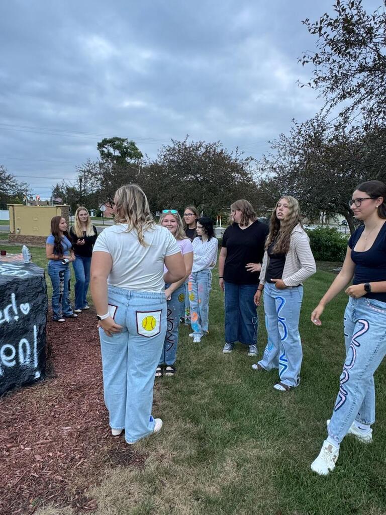 Group of young women in decorated jeans gather outdoors under a cloudy sky, chatting beside a colorful mural in a relaxed, friendly scene.
