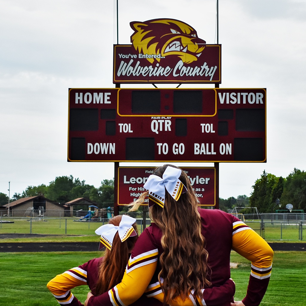 Two cheerleaders stand together arm in arm looking at the scoreboard. One wears a freshman bow and one wears a senior bow