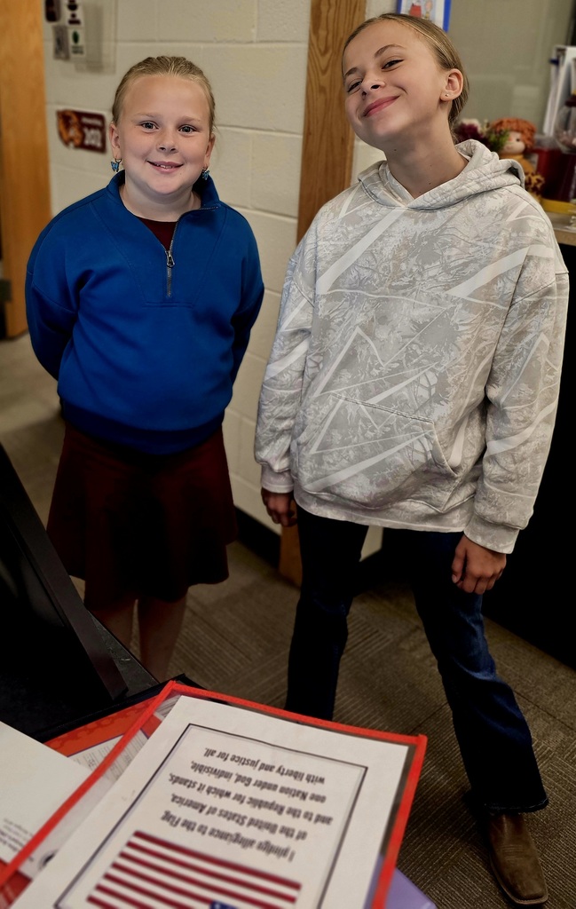Two 5th grade girls smiling in front of the microphone getting ready to leave the school in the Pledge of Allegiance. 