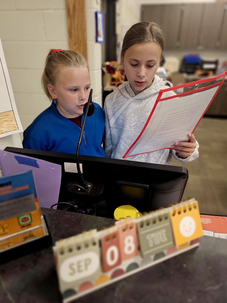 Two fifth grade girl students speak into a microphone leading the school in the Pledge of Allegiance. 