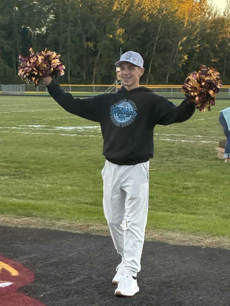 A high school boy in front of a football field holding pom poms and getting the crowd to cheer. 