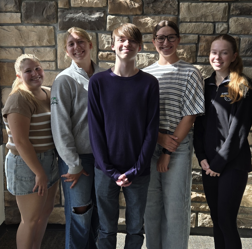 Five smiling teenagers pose together in sunlight against a stone wall, wearing casual clothes; the friendly group portrait conveys warmth and camaraderie.