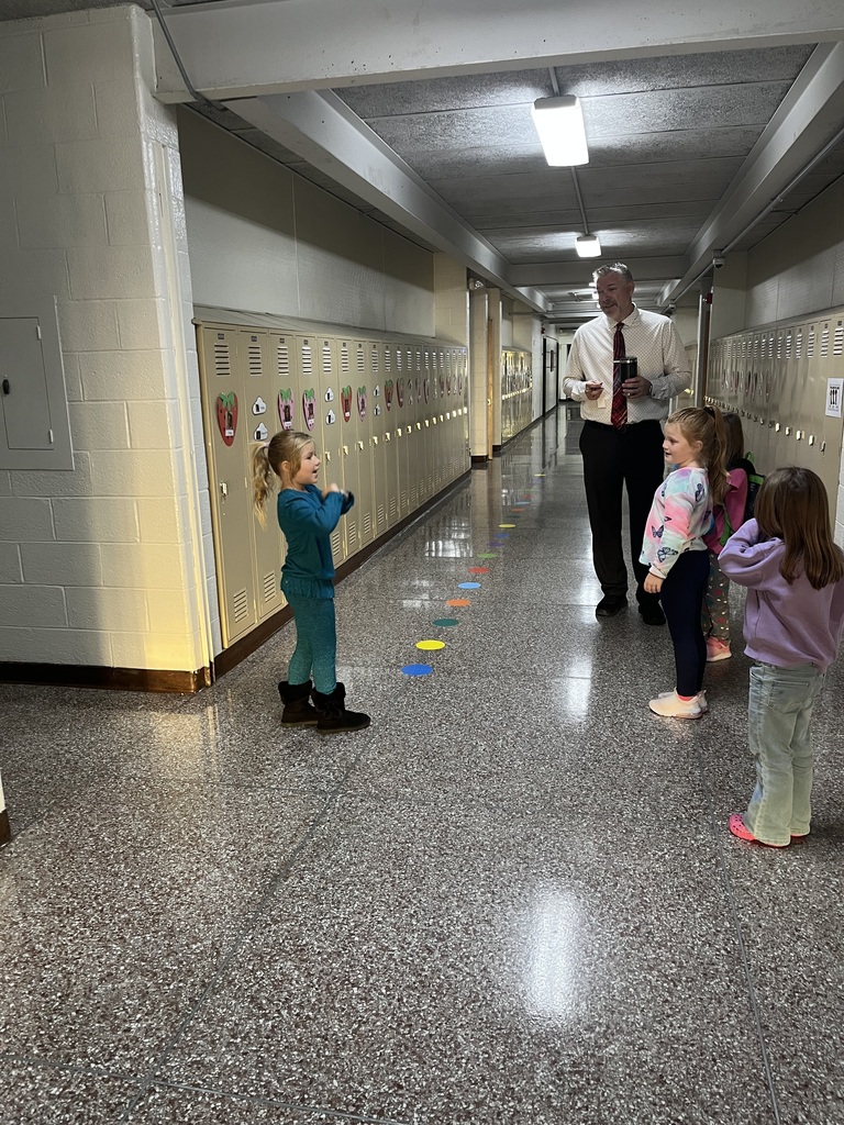 An educator and three girls gather in a school hallway beside colorful floor markers and decorated lockers; one girl gestures as others watch. The mood is warm and lively.