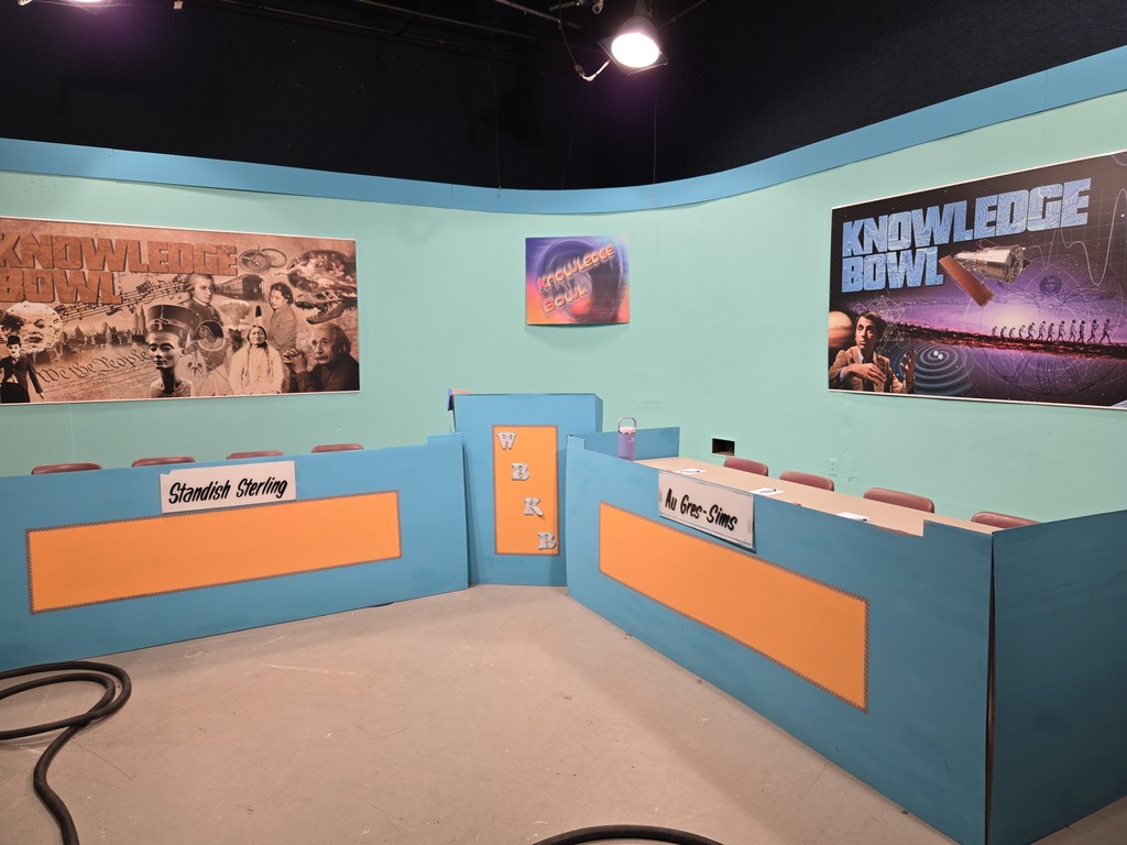 Two rows of blue and orange desks with name tags in front of a blue wall with three posters