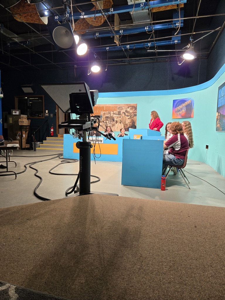Two people sitting at a blue desk in a room with a monitor, lights, and a carpeted floor.