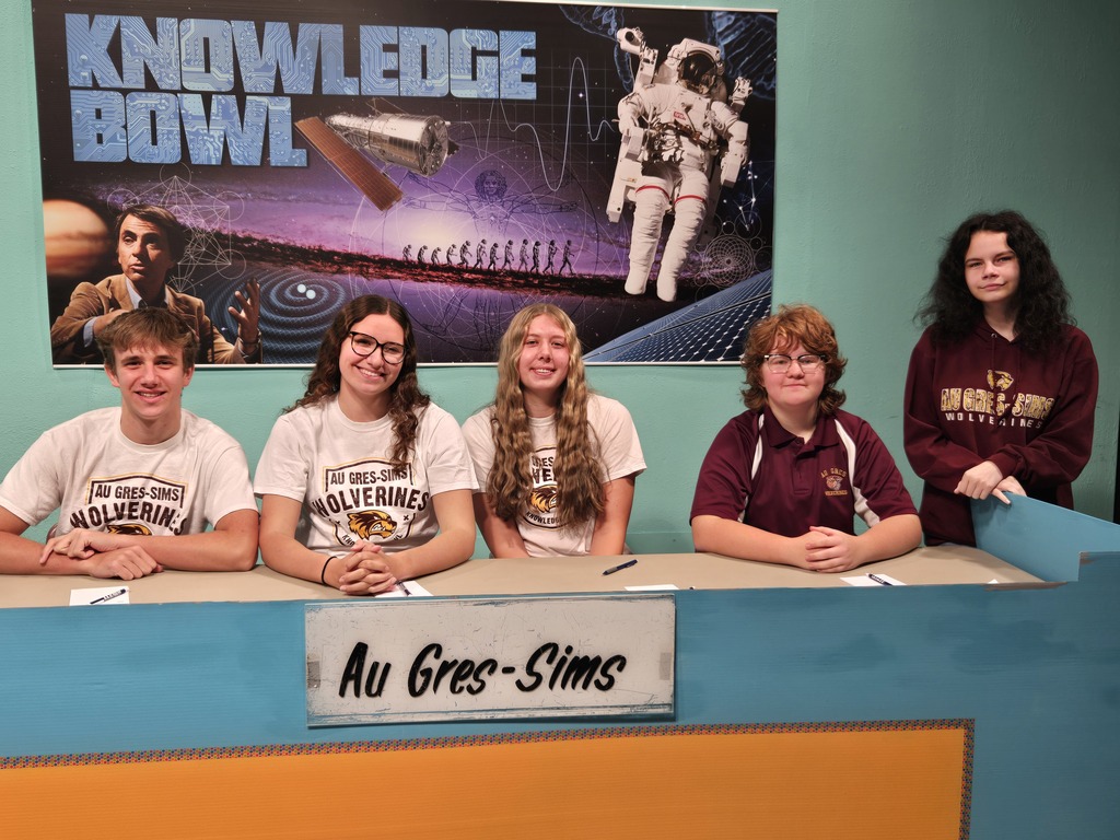 Five students sit at a table in front of a banner that says "Knowledge Bowl". Two students wear maroon shirts, three wear white.