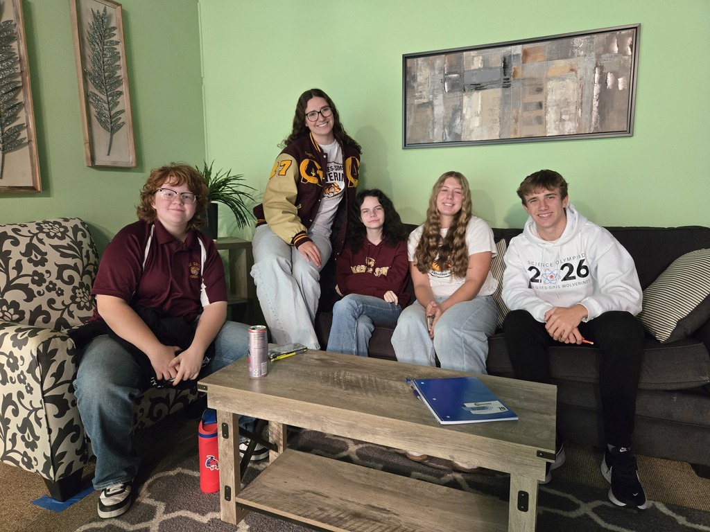 Six people sit on a couch and armchair. A table holds a book and a cup.