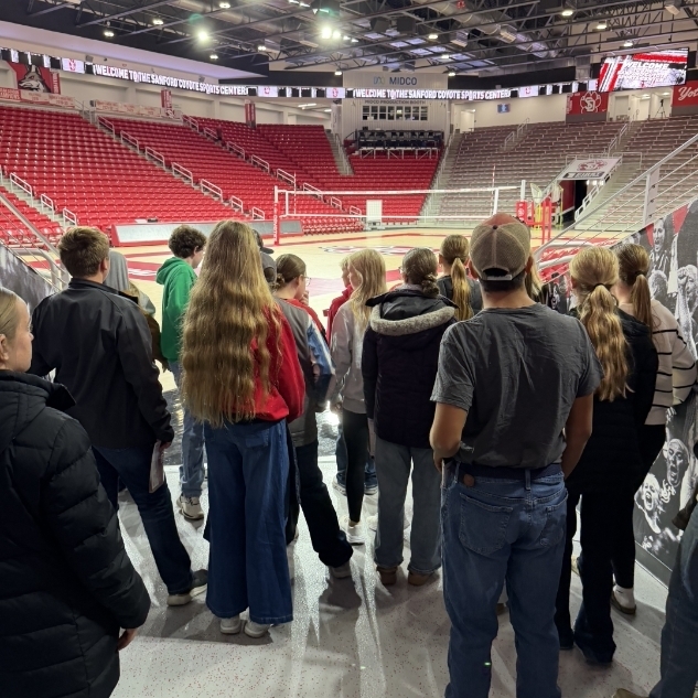 A group of students stands in a concourse area near a basketball court, facing a wall.