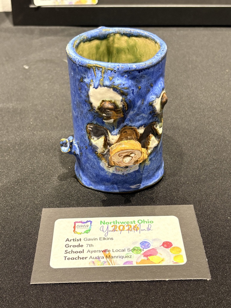 A blue ceramic mug created by Gavin Elkins is picture. The mug is modeled after a monster. It has unproportional eyes, sharp teeth, small hands, a tail handle, and is eating a gingerbread as half of it is hanging out of his mouth.