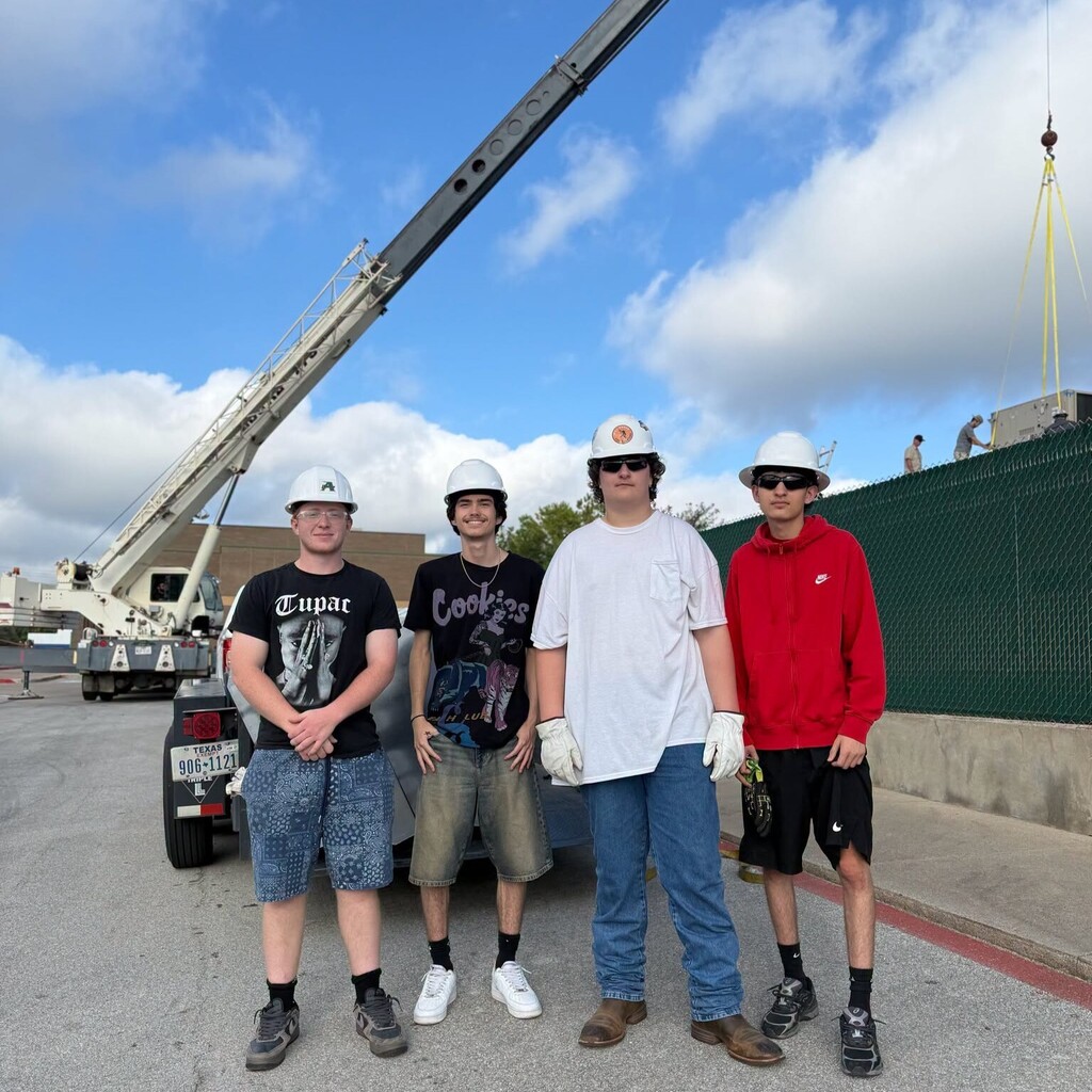four high school students wearing hard hats standing in front of crane