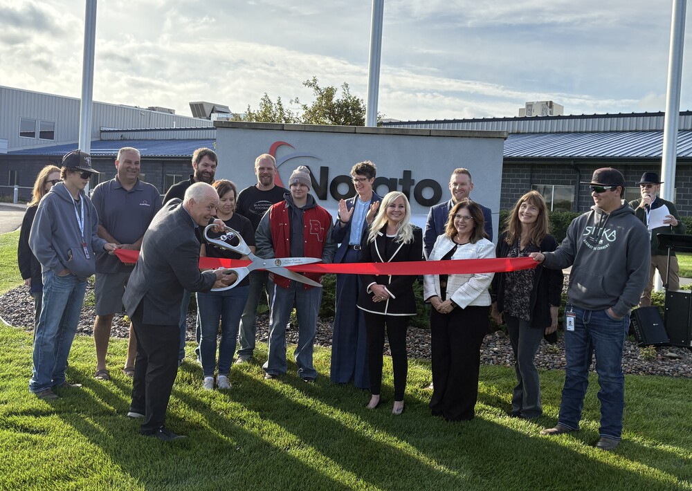 A group of people standing outdoors on a lawn for a ribbon-cutting ceremony in front of a company sign that reads "Nolato." On the left, a man in a dark suit jacket uses oversized silver scissors to cut a long red ribbon held across the front of the group. The attendees are smiling and watching, dressed in a mix of casual attire, hoodies, and business suits.