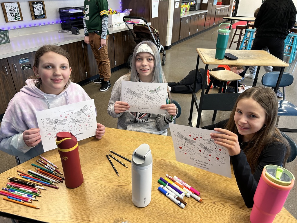 Three students at their desk holding up their artwork.