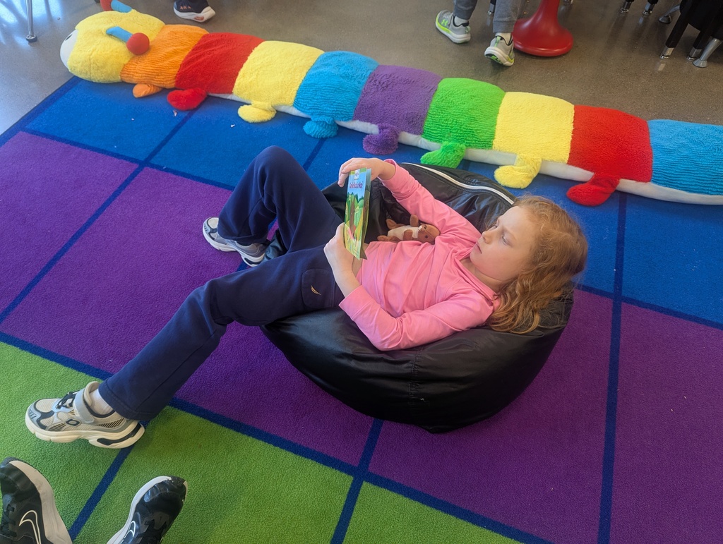 A little girl laying on a comfy chair reading and holding her stuffed animal.