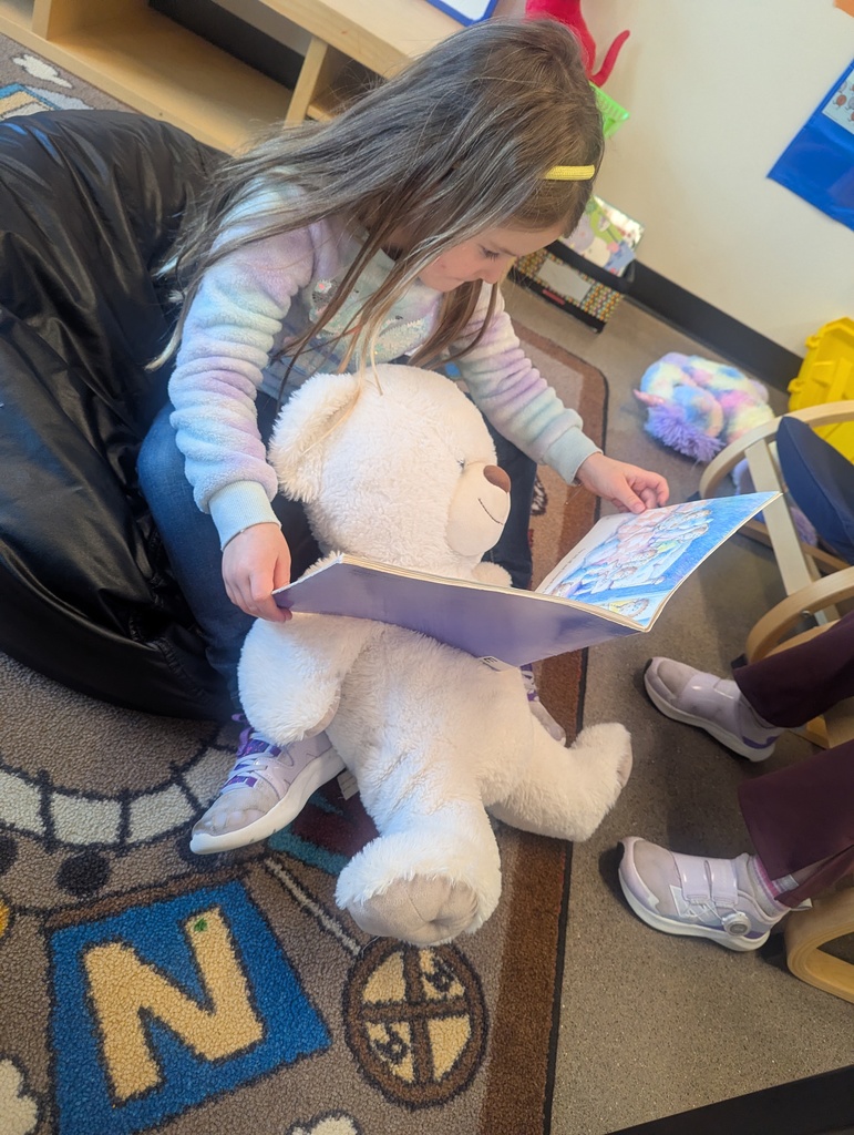 A little girl reading to her stuffed teddy bear.