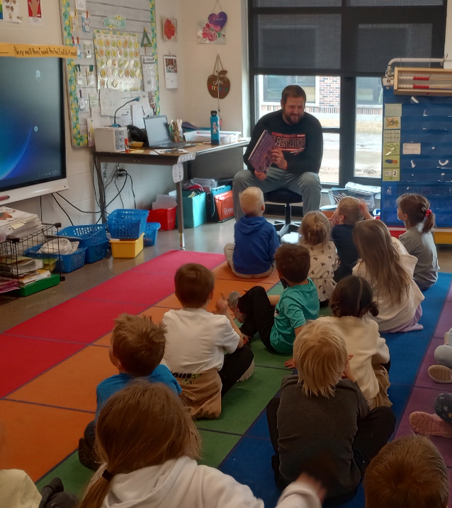 Principal Sbertoli reading a book to kindergartners sitting on the rug.