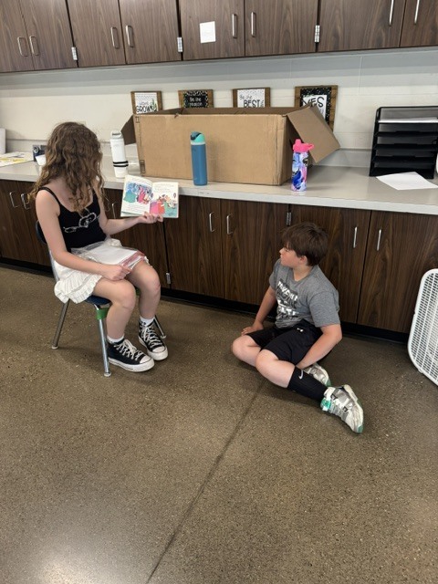 A student sitting in a chair reading a book to another student.