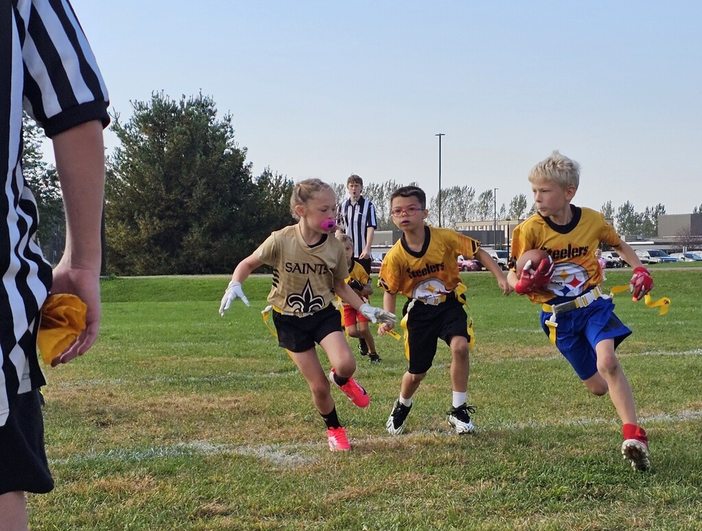 Children playing a game of youth flag football on a grassy field, with a ball carrier running past defenders.