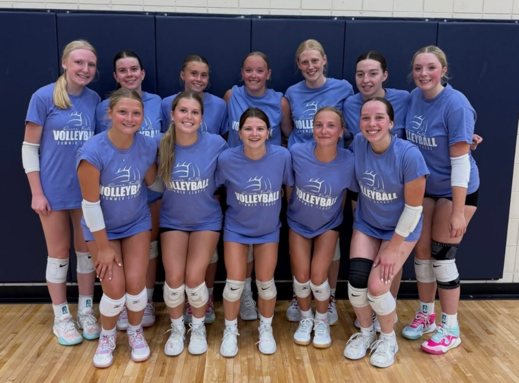 Group photo of a high school summer league volleyball team posing together in matching periwinkle blue t-shirts in a gymnasium.