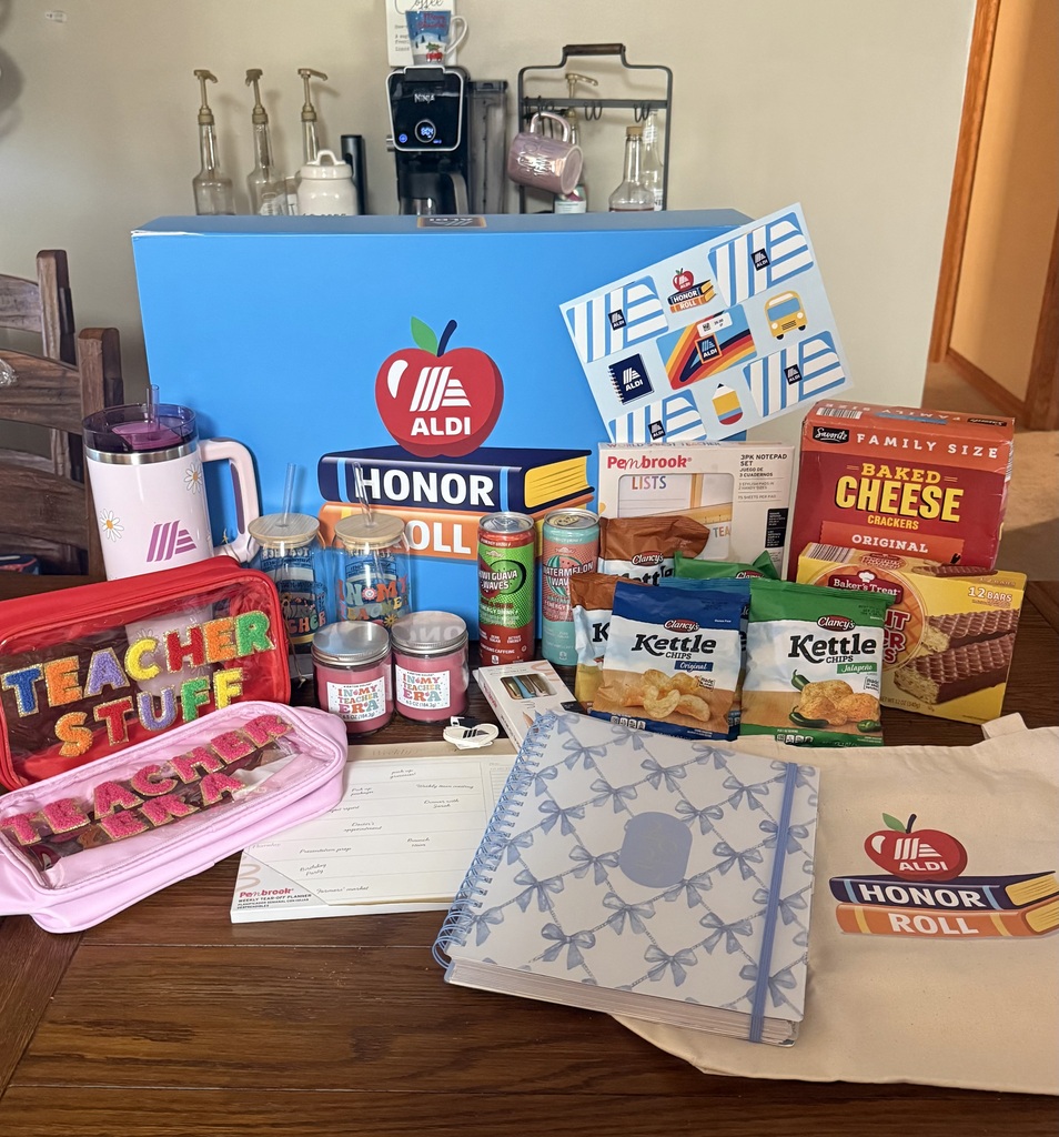 A wooden table filled with ALDI Teacher Honor Roll items, including a large blue box, school supplies, candles, tumblers, pouches, a tote bag, and various snacks like chips and crackers.