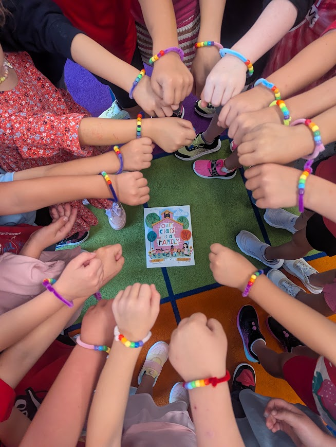 Overhead shot of kindergarten students standing in a circle with their hands reaching toward the center. Each child wears a colorful beaded bracelet made with pipe cleaners and rainbow-colored beads. In the middle of the circle on a colorful classroom rug lies the book "Our Class is a Family" by Shannon Olsen.