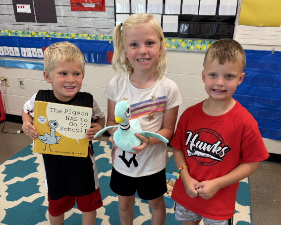 Three smiling kindergarten students stand inside a colorful classroom. The student on the left holds up the book "The Pigeon HAS to Go to School!", the middle student holds a stuffed Pigeon plush toy, and the student on the right wears a red Baldwin-Woodville Hawks shirt.
