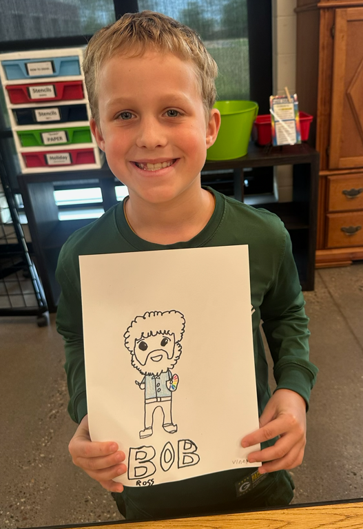 A young boy wearing a long-sleeved green shirt smiling proudly while holding up a drawing of a cartoon Bob Ross holding a paintbrush and color palette.