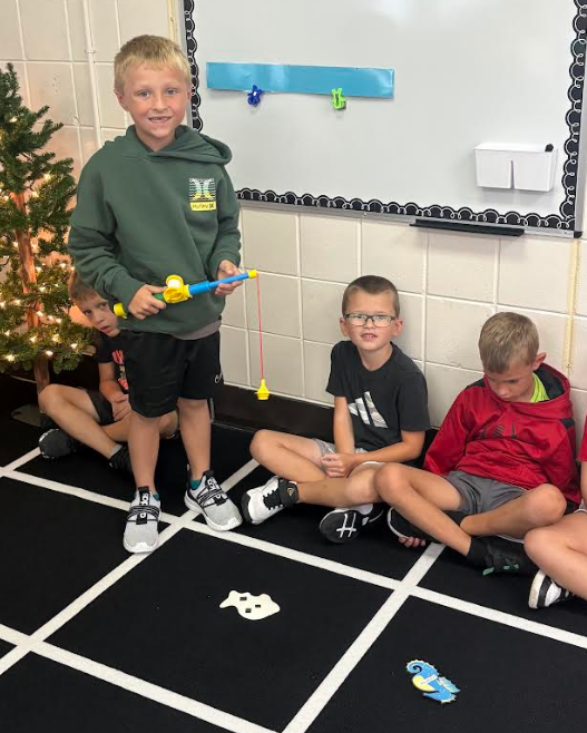 A young boy standing and holding a toy fishing pole over a black grid mat while three classmates sit nearby on the floor, practicing a "Fishing for School Rules" game in front of a whiteboard.