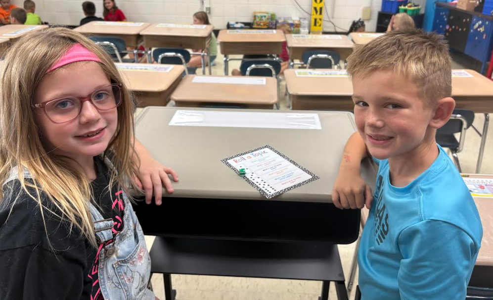 Two elementary students smiling together at a desk with a "Roll a Topic" icebreaker activity sheet placed in the center.