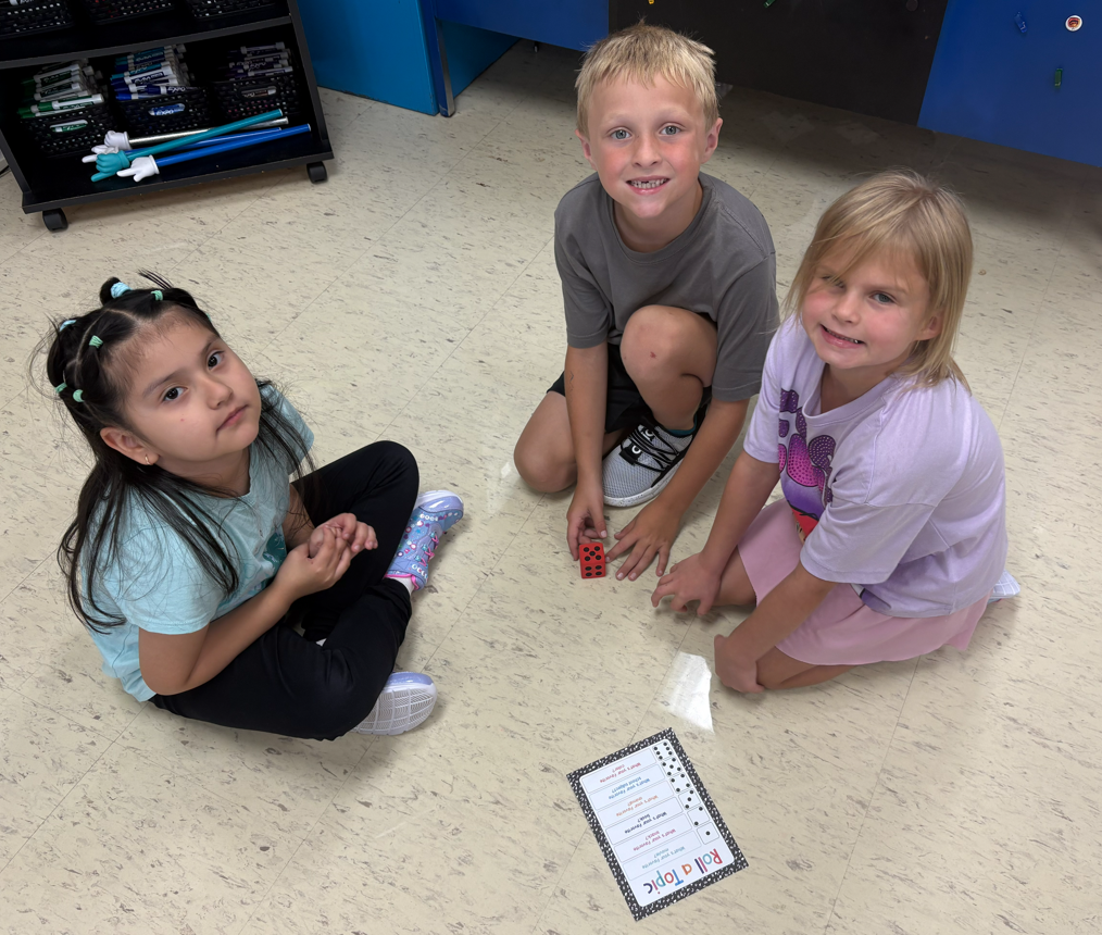Three elementary students sitting in a circle on the classroom floor around a red die and a printed "Roll a Topic" game card, smiling toward the camera.