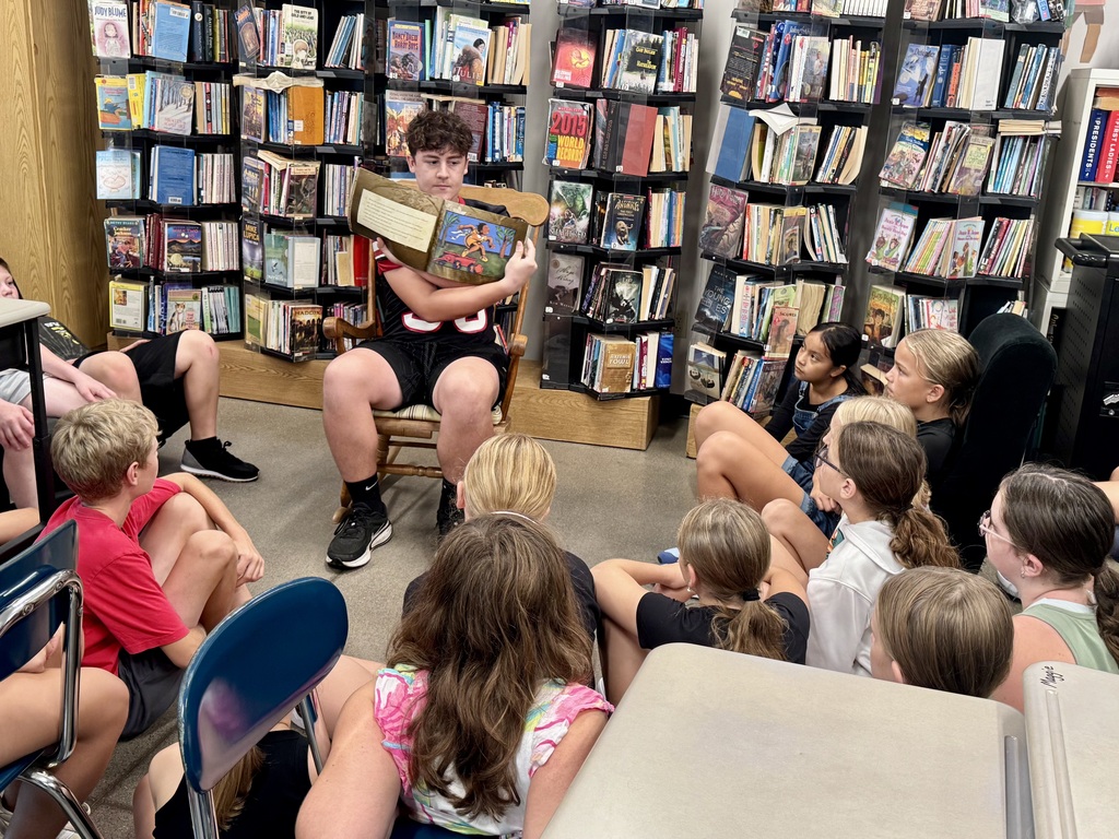 High school student Bryce sits in a wooden rocking chair in front of bookshelf-lined walls, holding open an illustrated picture book for a group of elementary students sitting in a semicircle around him on the floor.