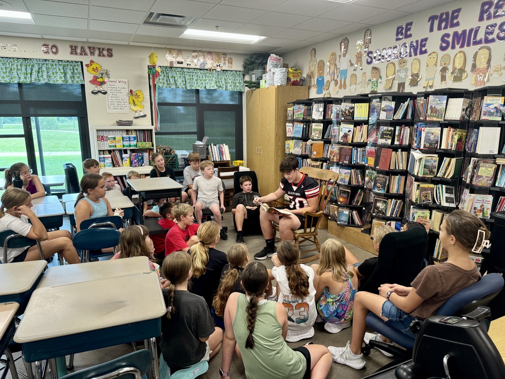 A wide shot of a classroom library area showing high school student Bryce reading to a circle of elementary students gathered around his rocking chair, surrounded by crowded bookshelves and classroom desks.